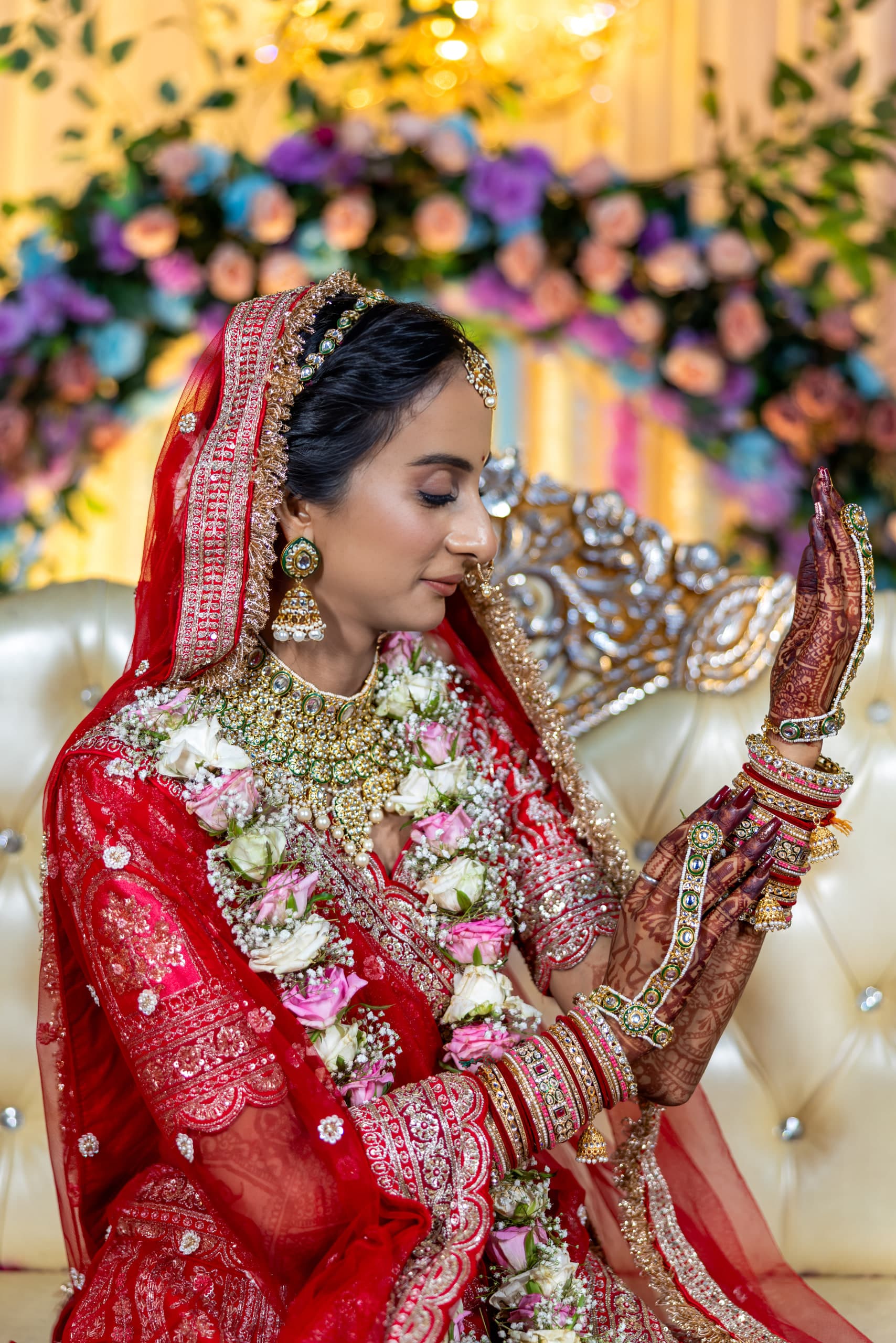 Seated bride turns to look at her own mehndi-covered hand, red and gold bangles stacked to the elbow