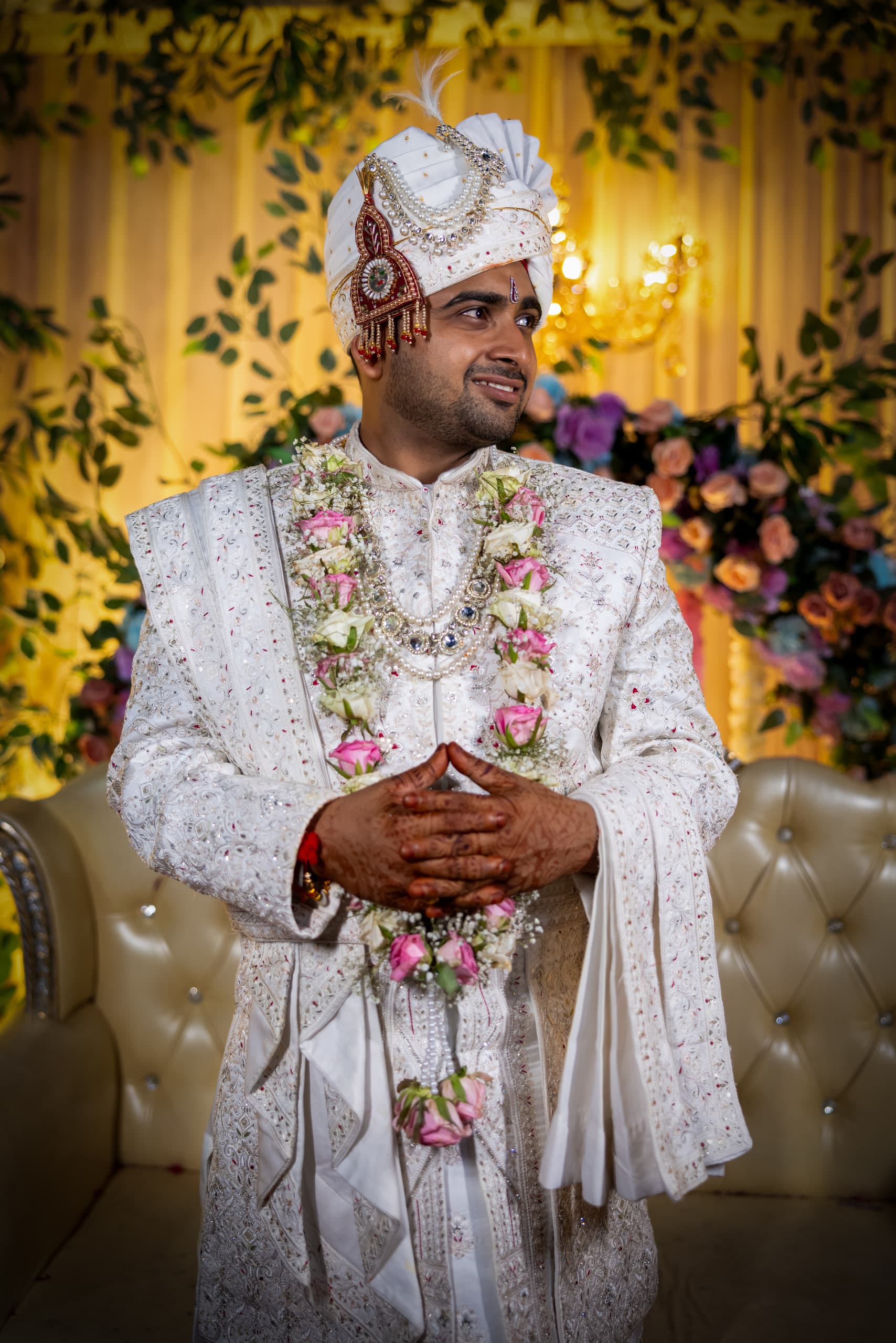 Groom in an ivory sherwani and safa looks off to one side, hands clasped, rose garland at his chest